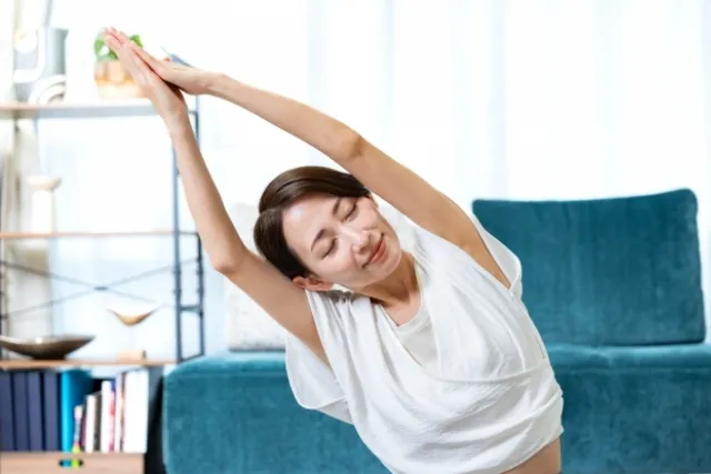 自宅の部屋で両掌を合わせて横にストレッチをしている白いウェアの女性