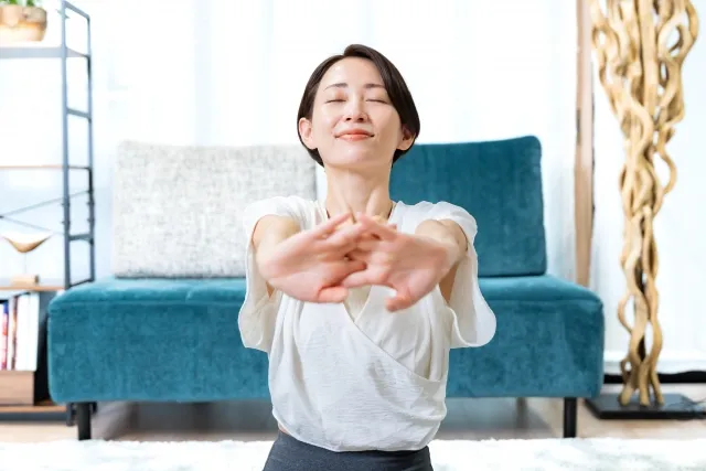 自宅の部屋で両手を組んで前に伸ばしている白いウェアの女性