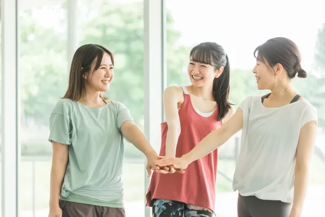 スタジオ内で微笑みながら円陣で片手を重ねる女性3人