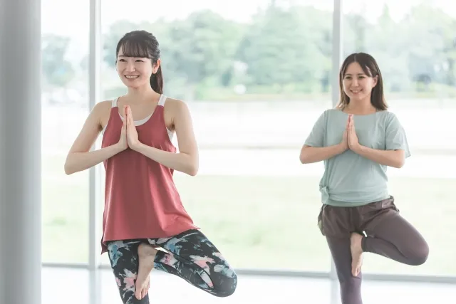 ヨガの木のポーズをしている赤いウェアの女性と薄い青色のウェアの女性