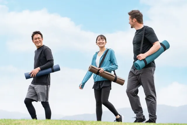 青空の下で巻いたヨガマットを肩で担いで歩きながら話をしている様子の男2名と女1名