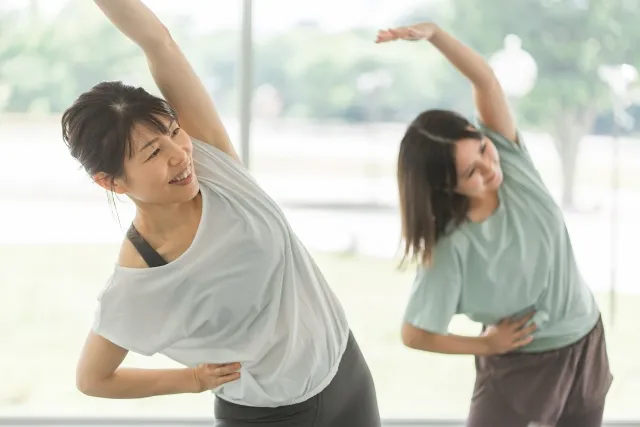 ピラティススタジオで片手を上げてストレッチをしている白いウェアの女性と緑のウェアの女性