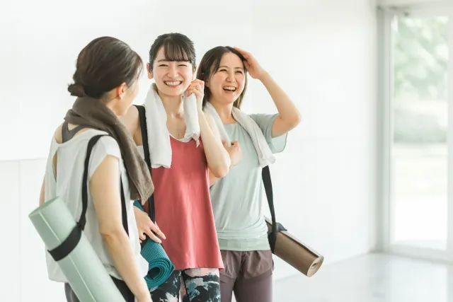 ピラティススタジオでタオルを首にかけストラップ付きのマットを肩から下げ笑顔の女性3人