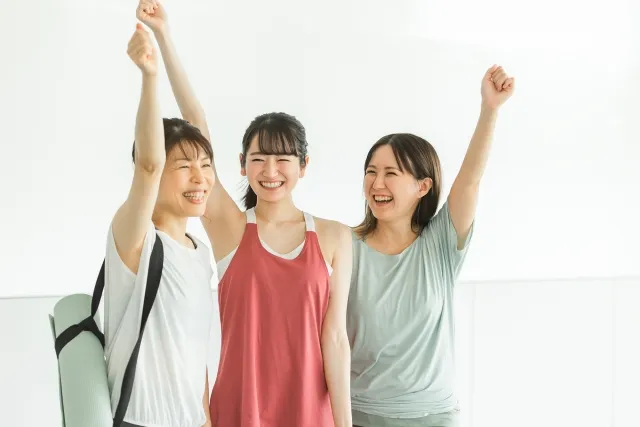 ピラティススタジオに通う3人の女性 それぞれ笑顔で拳を握って片手を上げている