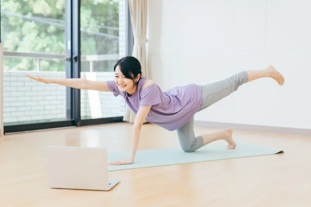 自宅でオンラインにてヨガの虎のポーズをしながら微笑んでいる紫色のウェアの女性