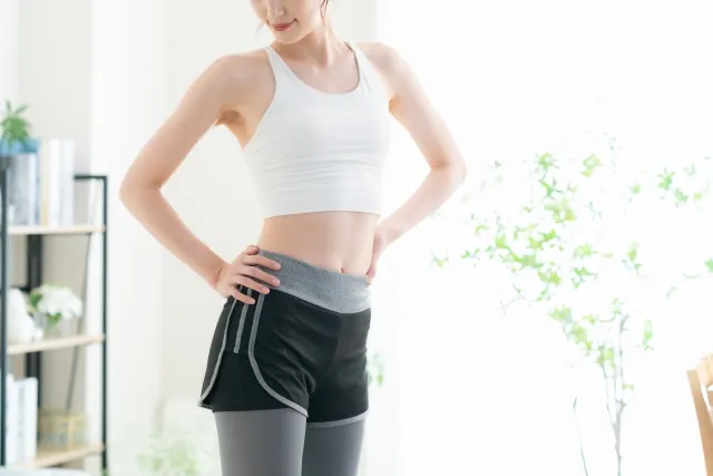 自宅で腰に両手を添えて腰回りを気にしている様子の細見の白いウェアの女性