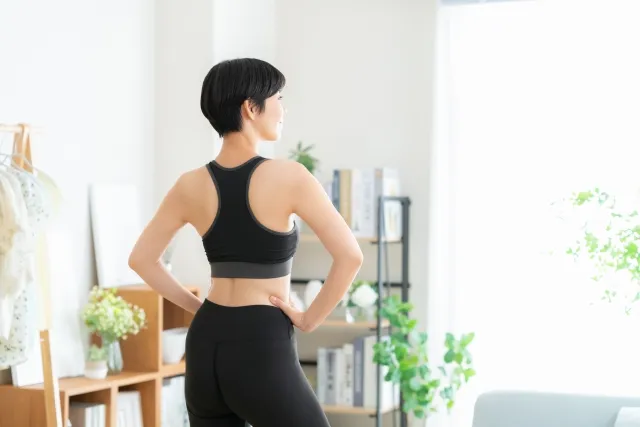 部屋で窓の方を向いて両手を腰にあてている黒色のウェアのショートカットの女性の後ろ姿