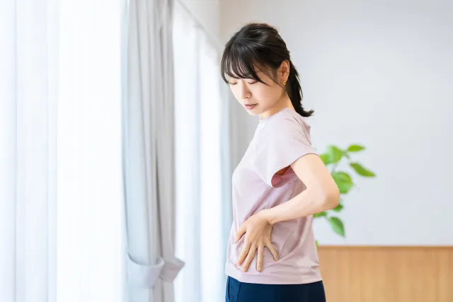 窓際で腰に手を当てて、くねらせた自身の腰を見ている薄紫色のウェアの女性