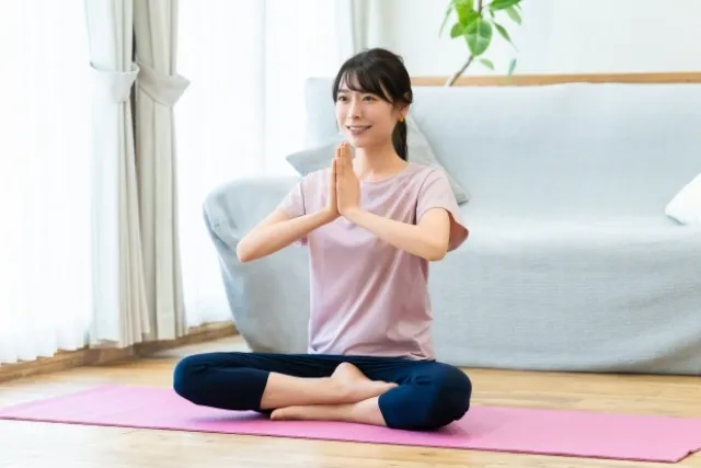 部屋のソファの前でピラティスマットの上で、胡坐をして合掌している薄紫色のウェアを着た女性