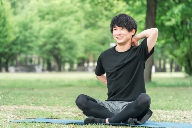 公園でヨガマットの上で胡坐をして背中の後ろで両手を上下で結ぼうとしている黒いウェアを着た男性