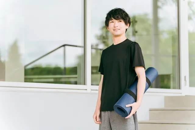 立ちながら丸めたヨガマットを片手で持っている黒いウェアの男性