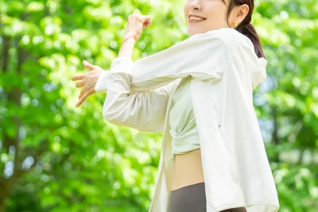 木々をバックにピラティスウェアを着て左手を右手の肘で伸ばしてストレッチをする女性