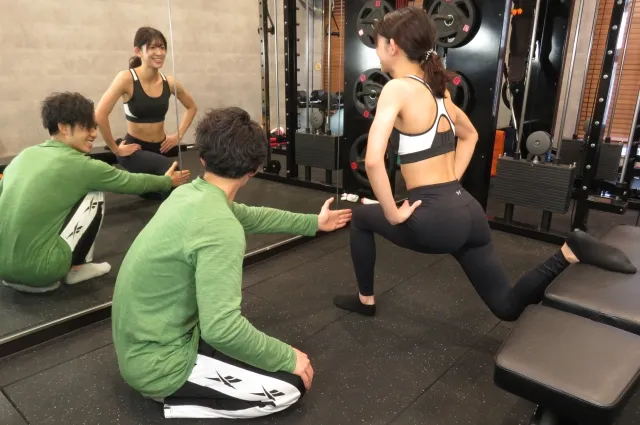 パーソナルジムARISANFITで鏡の前で器具を使って下半身のトレーニングをする女性と指導する男性トレーナー