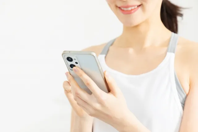 左手にスマホを持ち右手で操作しながら微笑む白いウェアを着た女性