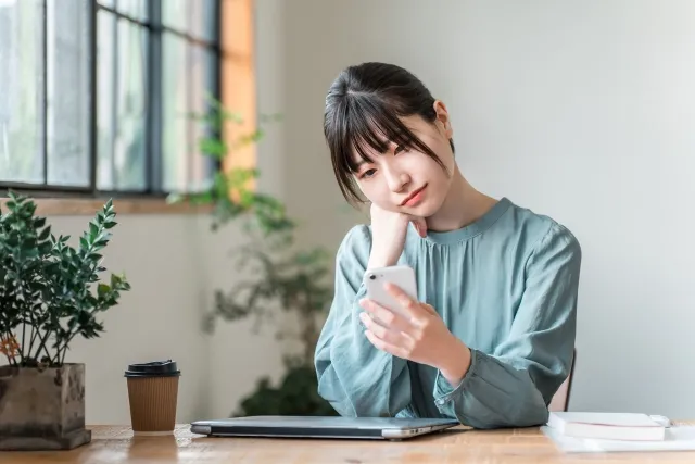 部屋でノートパソコンの置いてあるテーブルで頬杖をつきながらスマホを見ている女性