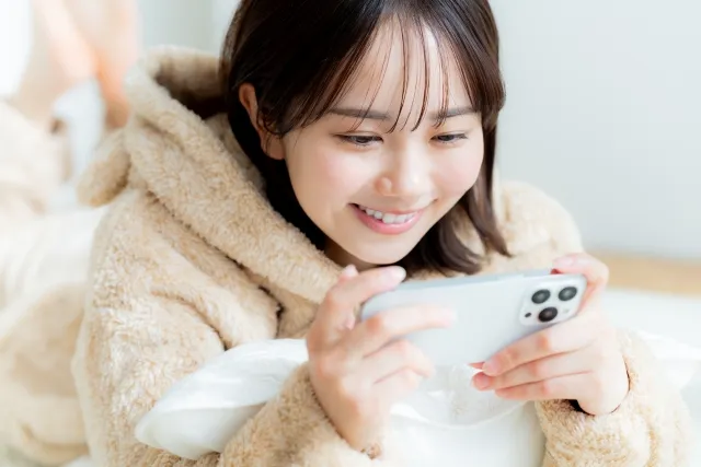 帽子に耳のついた茶色の動物の着ぐるみパジャマを着て寝転びながらスマホを見ている女性