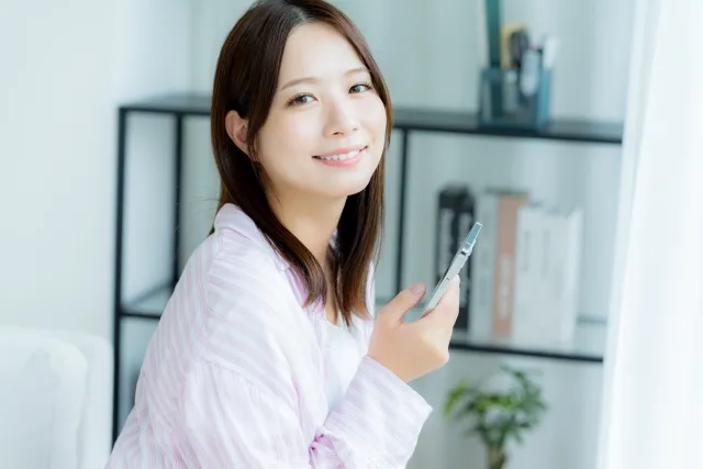 部屋で右手にスマホを持ちながら微笑む淡いピンク色のストライプのパジャマを着ている女性