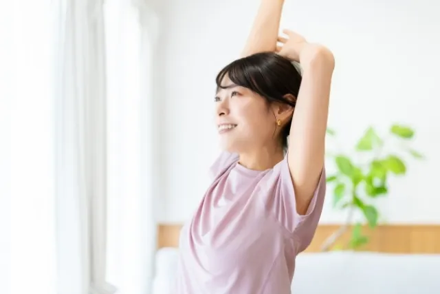 頭上に真っすぐ上げた右手の肘を左手で支えてストレッチする紫色のウェアの女性