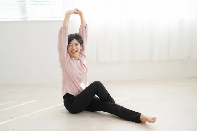 両手を組んで頭上に上げた状態で、左足は伸ばして、右足は膝を立てて左足を跨いだ状態で座わっている笑顔の女性