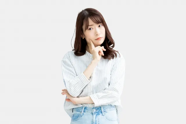 右手の人差し指を頬に当てながら考えているストライプのシャツにジーンズ姿の女性
