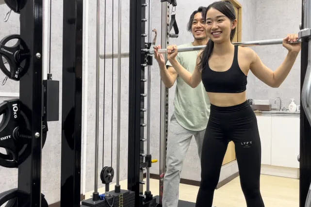パーソナルジムARISANFIT東高円寺店のトレーナーが立っている女性にトレーニング機材を使ってウエイトトレーニングの指導をしている