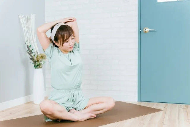 自宅にて胡坐して両手を頭の後ろで組んでストレッチしている女性