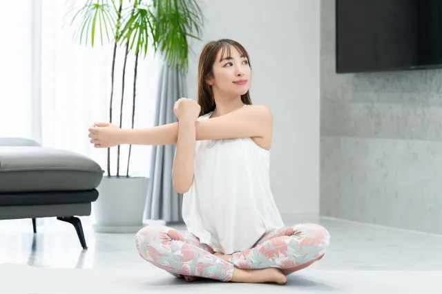 自宅の床に敷いたマットの上で胡坐をして伸ばした左手を右手で抑えてストレッチする女性
